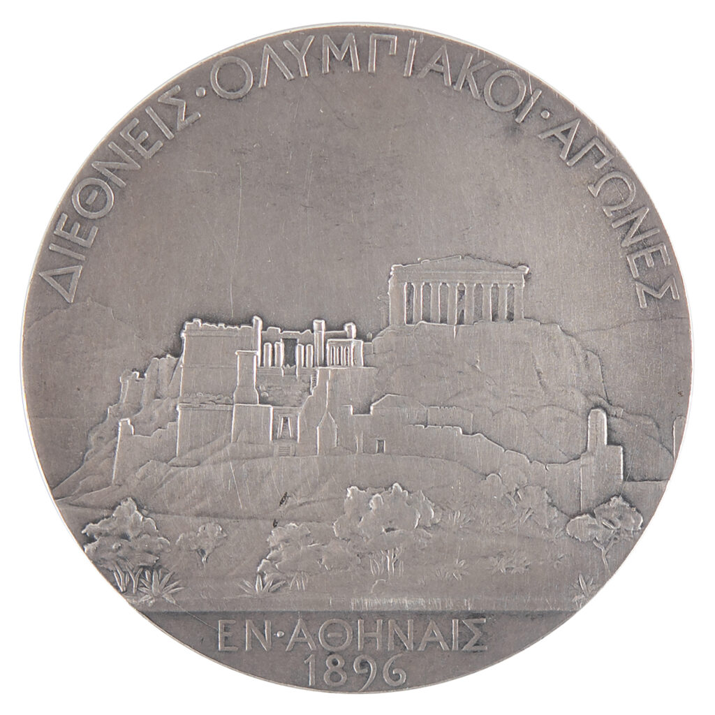 The reverse of the medal features a detailed view of the Acropolis of Athens topped by the Parthenon. The raised text to the upper and lower portion reads (translated), “International Olympic Games in Athens, 1896.”